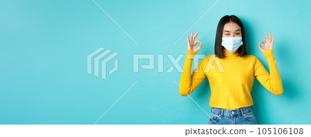 Covid-19, social distancing and pandemic concept. Cheerful asian girl in medical mask showing okay gestures, winking at camera, showing approval, standing over blue background Covid-19, social distancing and pandemic concept. Cheerful asian girl in medical mask showing okay gestures, winking at camera, showing approval, standing over blue background 105106108