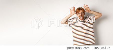 Funny young redhead man acting silly, showing tongue and mocking someone with stupid face, standing over white background 105106126