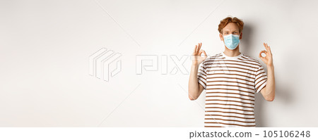 Covid-19, pandemic and social distancing concept. Young man with red hair wearing medical mask to prevent catching coronavirus, showing okay signs in approval, white background 105106248