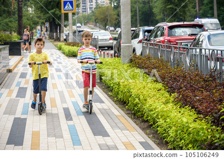 Two attractive European boys brothers, wearing red and white checkered shirts, standing on scooters in the park. They laughing, smiling, hugging and having fun. Active leisure time with kids Two attractive European boys brothers, wearing red and white checkered shirts, standing on scooters in the park. They laughing, smiling, hugging and having fun. Active leisure time with kids 105106249