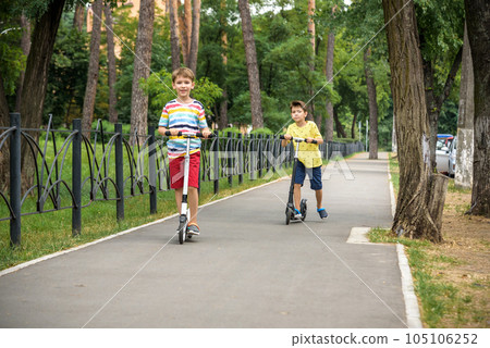 Two attractive European boys brothers, wearing red and white checkered shirts, standing on scooters in the park. They laughing, smiling, hugging and having fun. Active leisure time with kids 105106252