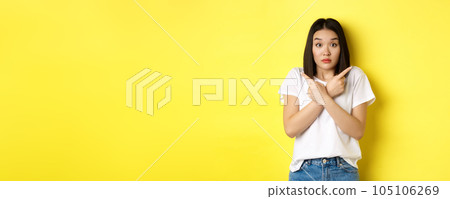 Indecisive asian girl need help with choice, pointing fingers sideways and looking confused, standing over yellow background 105106269