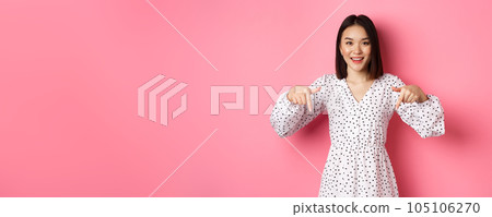 Look there. Cute asian woman in dress, pointing fingers down at copy space, showing product discount and smiling, standing over pink background 105106270