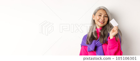 Shopping concept. Stylish asian senior woman smiling and showing plastic credit card, paying contactless, standing over white background 105106301
