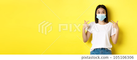 Covid-19, quarantine and social distancing concept. Young asian woman in white t-shirt, pointing at her medical mask from coronavirus, yellow background Covid-19, quarantine and social distancing concept. Young asian woman in white t-shirt, pointing at her medical mask from coronavirus, yellow background 105106306