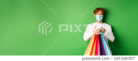 Pandemic and lifestyle concept. Cheerful redhead man going shopping in store, wearing medical mask and holding bags, standing over green background Pandemic and lifestyle concept. Cheerful redhead man going shopping in store, wearing medical mask and holding bags, standing over green background 105106307