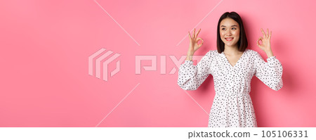 Pretty young asian woman in dress showing okay sign, praising and showing approval, looking satisfied, standing against pink background Pretty young asian woman in dress showing okay sign, praising and showing approval, looking satisfied, standing against pink background 105106331