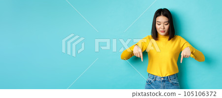 Shopping concept. Young teenage korean girl pointing and looking down at commercial, standing against blue background Shopping concept. Young teenage korean girl pointing and looking down at commercial, standing against blue background 105106372