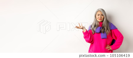 Angry and confused asian senior woman spread hand sideways and staring at camera puzzled, standing in pink sweater against white background Angry and confused asian senior woman spread hand sideways and staring at camera puzzled, standing in pink sweater against white background 105106419