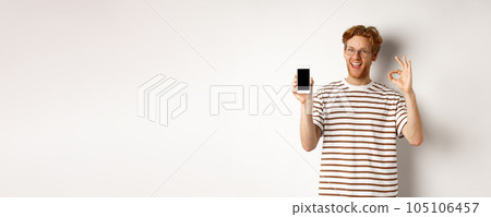 Technology and e-commerce concept. Young man with red hair showing okay sign and blank smartphone screen, praising awesome app, standing over white background Technology and e-commerce concept. Young man with red hair showing okay sign and blank smartphone screen, praising awesome app, standing over white background 105106457