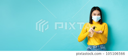 Covid-19, social distancing and pandemic concept. Smiling asian woman in medical mask using preventive measures from coronavirus, using hand sanitizer, standing over blue background 105106566