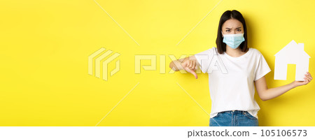 Covid-19 and real estate concept. Disappointed asian woman in medical mask, showing thumb down and paper house cutout, standing upset against yellow background Covid-19 and real estate concept. Disappointed asian woman in medical mask, showing thumb down and paper house cutout, standing upset against yellow background 105106573