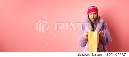 Upset senior asian woman open shopping bag and looking sad at camera, standing over pink background Upset senior asian woman open shopping bag and looking sad at camera, standing over pink background 105106587