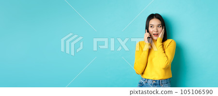 Woman looking intrigued while talking on mobile phone, receive interesting offer during call, standing over blue background 105106590