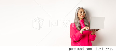 Senior asian woman working freelance, using laptop and smiling, standing over white background 105106649