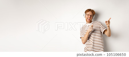 Technology concept. Happy redhead man listening music in headphones and dancing and pointing fingers right, standing over white background 105106650