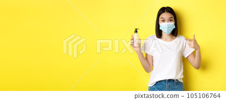 Covid, health care and pandemic concept. Asian girl in face mask from coronavirus, pointing finger at good hand sanitizer, standing over yellow background 105106764