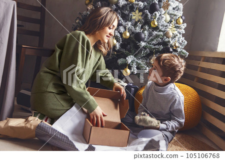 Little boy unpack Christmas gift with mom while sitting near xmas tree, kid open New Year present 105106768