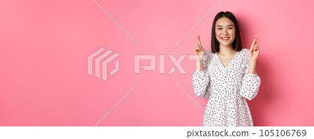 Cute asian woman making a wish, smiling happy and cross fingers for good luck, waiting over pink background 105106769
