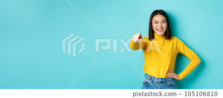 Beauty and fashion concept. Cheerful asian woman laughing and smiling, pointing finger at camera, choose you, standing over blue background 105106810