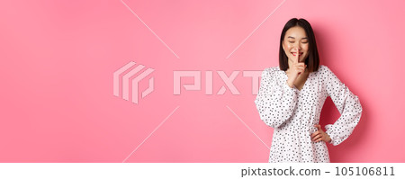 Beautiful Korean woman in trendy dress asking to keep secret, hushing with soft smile and closed eyes, standing over pink background 105106811