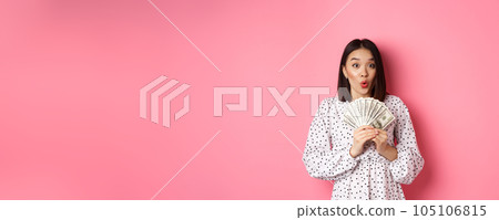 Shopping concept. Excited asian woman holding money, gasping amazed and staring at camera, standing over pink background 105106815
