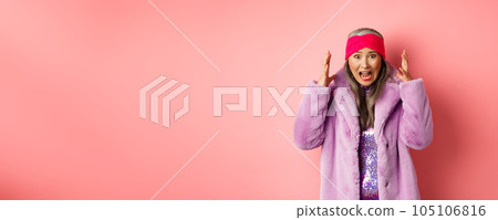 Anxious asian senior woman in stylish faux fur coat screaming, shaking hands and shouting alarmed and worried at camera, standing over pink background 105106816