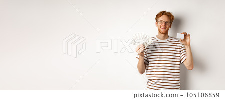 Shopping and finance concept. Young redhead man with beard and glasses showing plastic credit card with money in dollars, white background Shopping and finance concept. Young redhead man with beard and glasses showing plastic credit card with money in dollars, white background 105106859