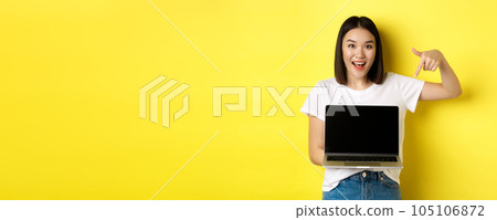 Young asian woman demonstrate online offer, showing laptop screen and smiling, standing over yellow background 105106872