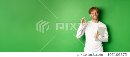 Handsome young male employee in glasses showing Ok sign, holding laptop, standing over green background Handsome young male employee in glasses showing Ok sign, holding laptop, standing over green background 105106875