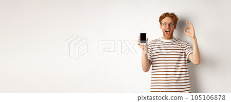 Technology and e-commerce concept. Young man with red hair showing okay sign and blank smartphone screen, praising awesome app, standing over white background Technology and e-commerce concept. Young man with red hair showing okay sign and blank smartphone screen, praising awesome app, standing over white background 105106878