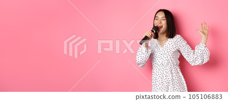 Happy asian woman dancing and singing in microphone, performing at karaoke, standing over pink background Happy asian woman dancing and singing in microphone, performing at karaoke, standing over pink background 105106883