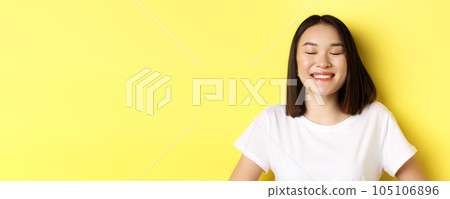 Close up of happy, romantic asian girl dreaming of something, close eyes and smiling delighted, standing over yellow background Close up of happy, romantic asian girl dreaming of something, close eyes and smiling delighted, standing over yellow background 105106896