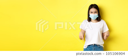 Covid-19, pandemic and social distancing concept. Disappointed asian girl in medical mask, frowning upset and pointing fingers down at logo, standing over yellow background Covid-19, pandemic and social distancing concept. Disappointed asian girl in medical mask, frowning upset and pointing fingers down at logo, standing over yellow background 105106904