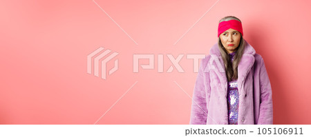 Sad and indecisive asian senior woman in fashionable faux fur coat looking left and sulking upset, standing against pink background 105106911