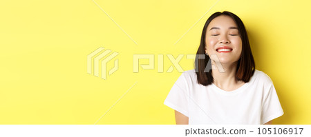Close up of happy and relaxed asian woman enjoying sun, smiling with eyes closed and looking joyful, standing over yellow background Close up of happy and relaxed asian woman enjoying sun, smiling with eyes closed and looking joyful, standing over yellow background 105106917