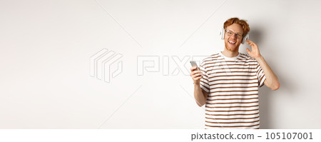 Technology concept. Young man with red hair and beard listening music in headphones and using smartphone, smiling at camera, white background Technology concept. Young man with red hair and beard listening music in headphones and using smartphone, smiling at camera, white background 105107001