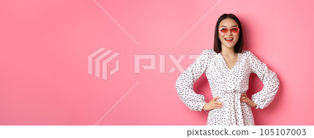 Tourism and lifestyle concept. Beautiful korean woman on vacation, wearing sunglasses and dress, looking left with happy smile, standing over pink background Tourism and lifestyle concept. Beautiful korean woman on vacation, wearing sunglasses and dress, looking left with happy smile, standing over pink background 105107003