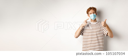 Covid-19, pandemic and social distancing concept. Cheerful young man with red hair pointing at medical mask and showing thumb-up, recommend use preventive measures against virus Covid-19, pandemic and social distancing concept. Cheerful young man with red hair pointing at medical mask and showing thumb-up, recommend use preventive measures against virus 105107034