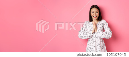 Beautiful asian woman thanking you, holding hands together in appreciation gesture, smiling happy at camera, standing grateful over pink background 105107045