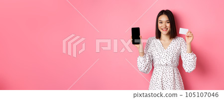 Cute asian woman shopping online, showing bank credit card and mobile screen, smiling and looking at camera, standing over pink background 105107046
