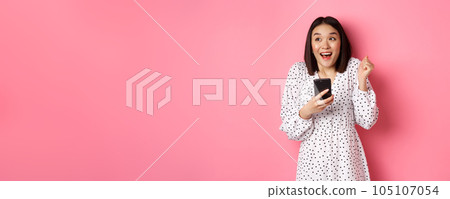 Online shopping and beauty concept. Excited asian woman winning in internet, holding smartphone and rejoicing, smiling happy and celebrating, standing over pink background Online shopping and beauty concept. Excited asian woman winning in internet, holding smartphone and rejoicing, smiling happy and celebrating, standing over pink background 105107054