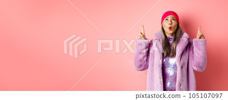Impressed asian elderly woman in stylish fake-fur coat checking out promo, looking and pointing fingers up, standing over pink background Impressed asian elderly woman in stylish fake-fur coat checking out promo, looking and pointing fingers up, standing over pink background 105107097