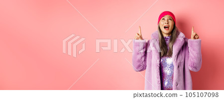 Mature asian hipster woman pointing fingers up, looking at top promo with amazement, checking out special deal, standing over pink background 105107098