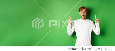 Shocked and worried redhead man in glasses gasping and staring at camera, pointing fingers up at advertisement, standing over green background 105107099