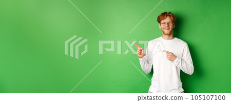 Handsome young man with red hair and beard smiling, pointing fingers left at logo, showing advertisement, standing over green background Handsome young man with red hair and beard smiling, pointing fingers left at logo, showing advertisement, standing over green background 105107100