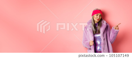 Trendy asian senior woman in cool purple winter coat pointing finger right, showing advertisement and looking sassy at camera, pink background Trendy asian senior woman in cool purple winter coat pointing finger right, showing advertisement and looking sassy at camera, pink background 105107149