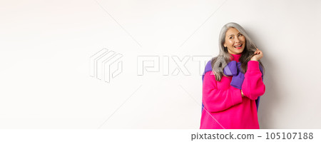 Valentines day concept. Beautiful asian senior woman laughing coquettish, playing with hair strands and looking at upper left corner flirty, standing over white background 105107188