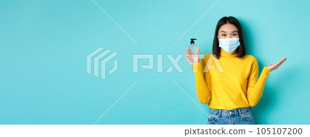 Covid-19, social distancing and pandemic concept. Cheerful asian woman showing hand sanitizer, recommend use antiseptic from coronavirus, wearing medical mask 105107200