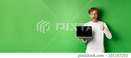 Impressed caucasian man with red hair and beard, showing blank laptop screen and thumb-up, looking amazed at camera, praising something online, green background 105107203